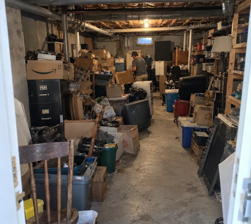 A basement lined with boxes, storage bins, filing cabinets, an old TV and furniture, a crew member at the back