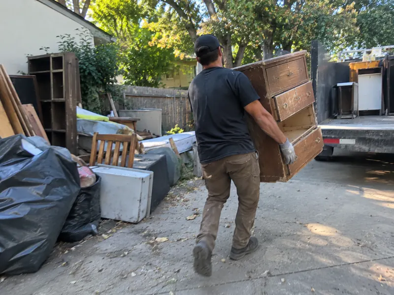 A crew member carrying an old wooden dresser down a driveway to the truck, past bags, a bookcase and more old furniture