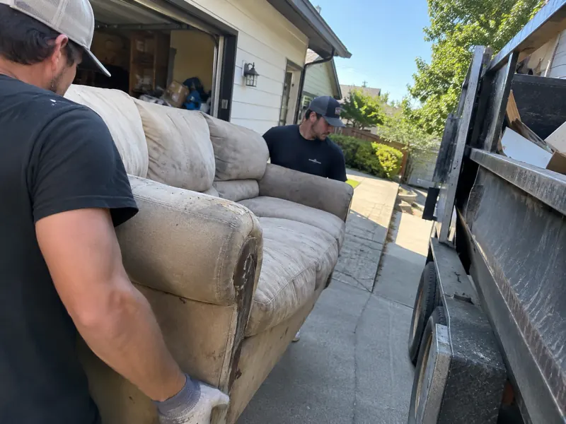 Two crew members carrying a worn couch out of a garage and down the driveway on a junk hauling job