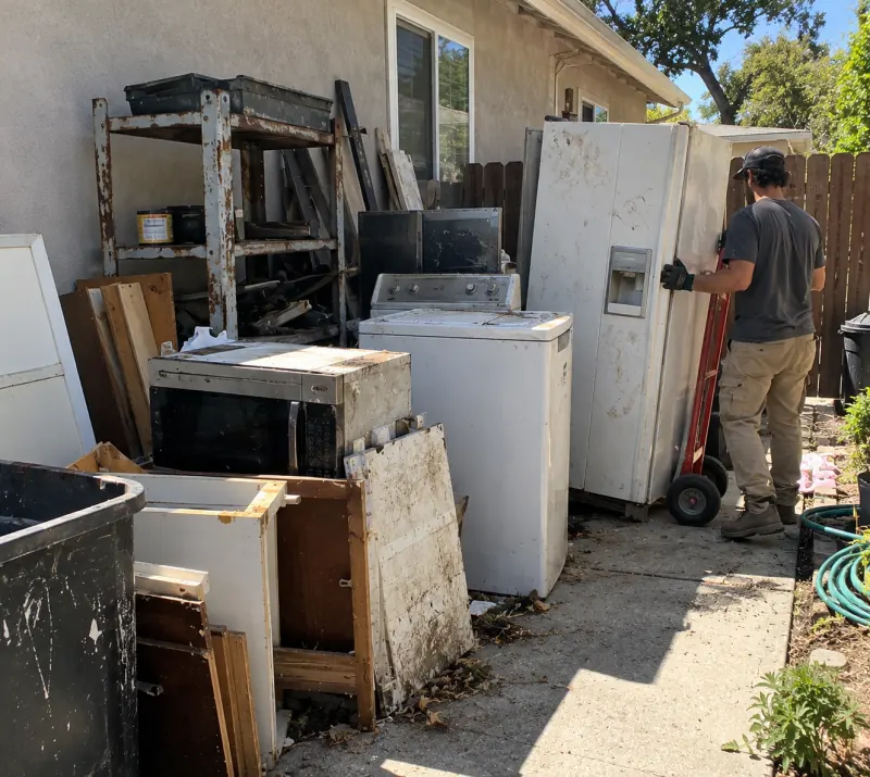 A crew member wheeling an old side-by-side fridge on a hand truck past a washer, a microwave and rusted shelving