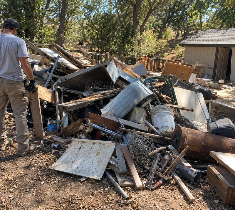 A crew member working through a pile of old lumber, sheet metal, wire fencing, buckets and a rusted tank beside a shed