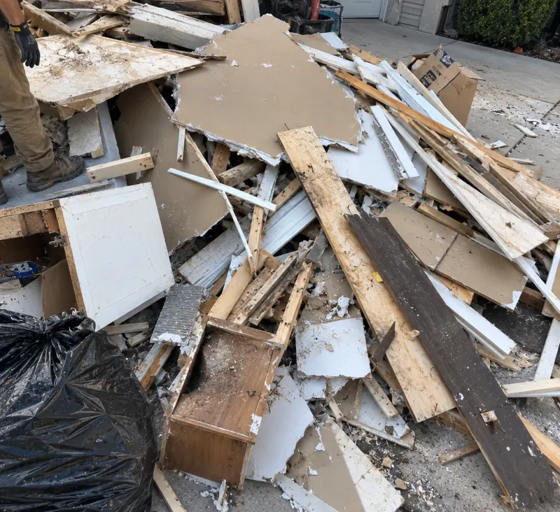 A driveway pile of broken drywall, boards, trim and an old cabinet box, with a crew member at the edge
