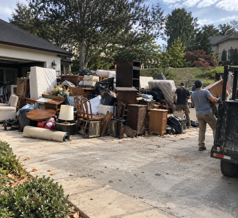 A driveway covered with dressers, chairs, mattresses, a table, lamps and bags while two crew members load a trailer