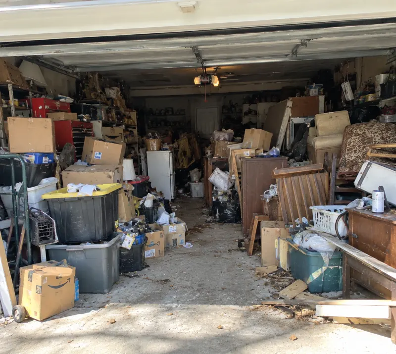 An open garage packed to the back wall with boxes, storage bins, a small fridge, chairs and dressers, before a cleanout