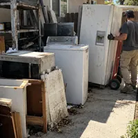 A crew member moving an old fridge on a hand truck past a washer