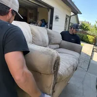 Two crew members carrying a worn couch out of a garage