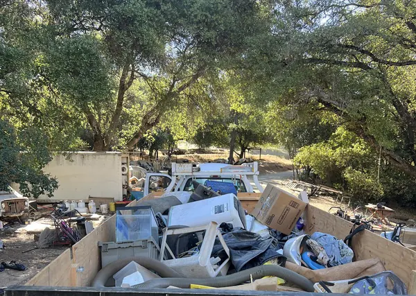 An open trailer packed with boxes, old appliances, bags and furniture under oak trees