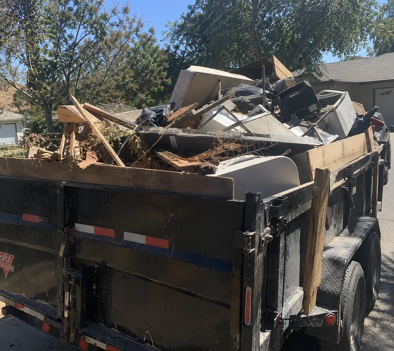 A black dump trailer loaded above its sides with lumber, branches, bags, a mattress and household junk