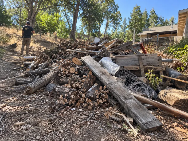 Cut logs, weathered boards, wire fencing and dry brush piled on a hillside lot under pines and oaks, a crew member raking